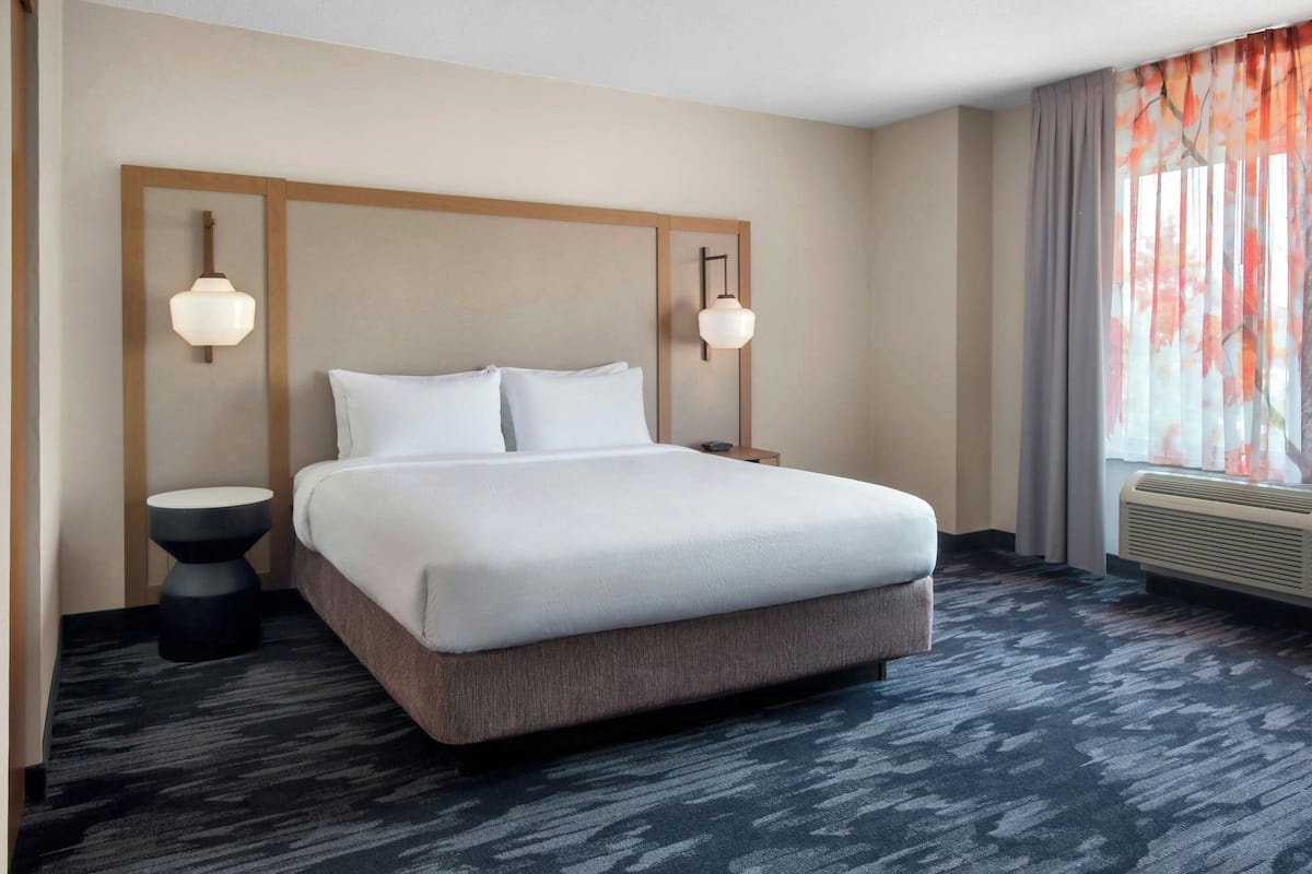 A hotel room with a large bed, two bedside tables, a wall-mounted light fixture, and a window with curtains.