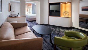 A modern hotel room with a sofa, a small table, a green chair, and a TV mounted on the wall. - Fairfield Inn & Suites by Marriott Woodbridge (Avenel)