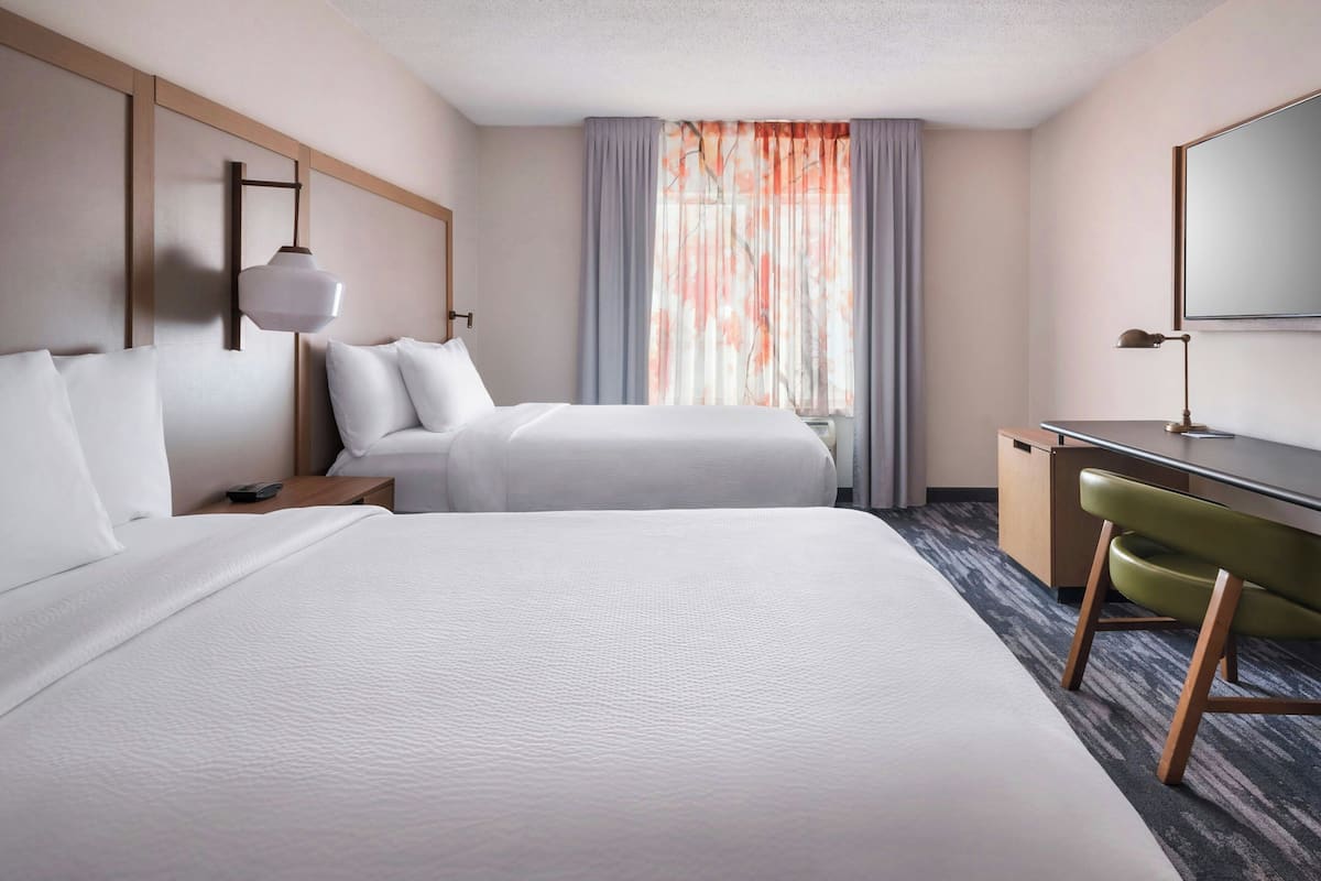 A hotel room with two beds, a desk, a chair, a lamp, and a large window with curtains.