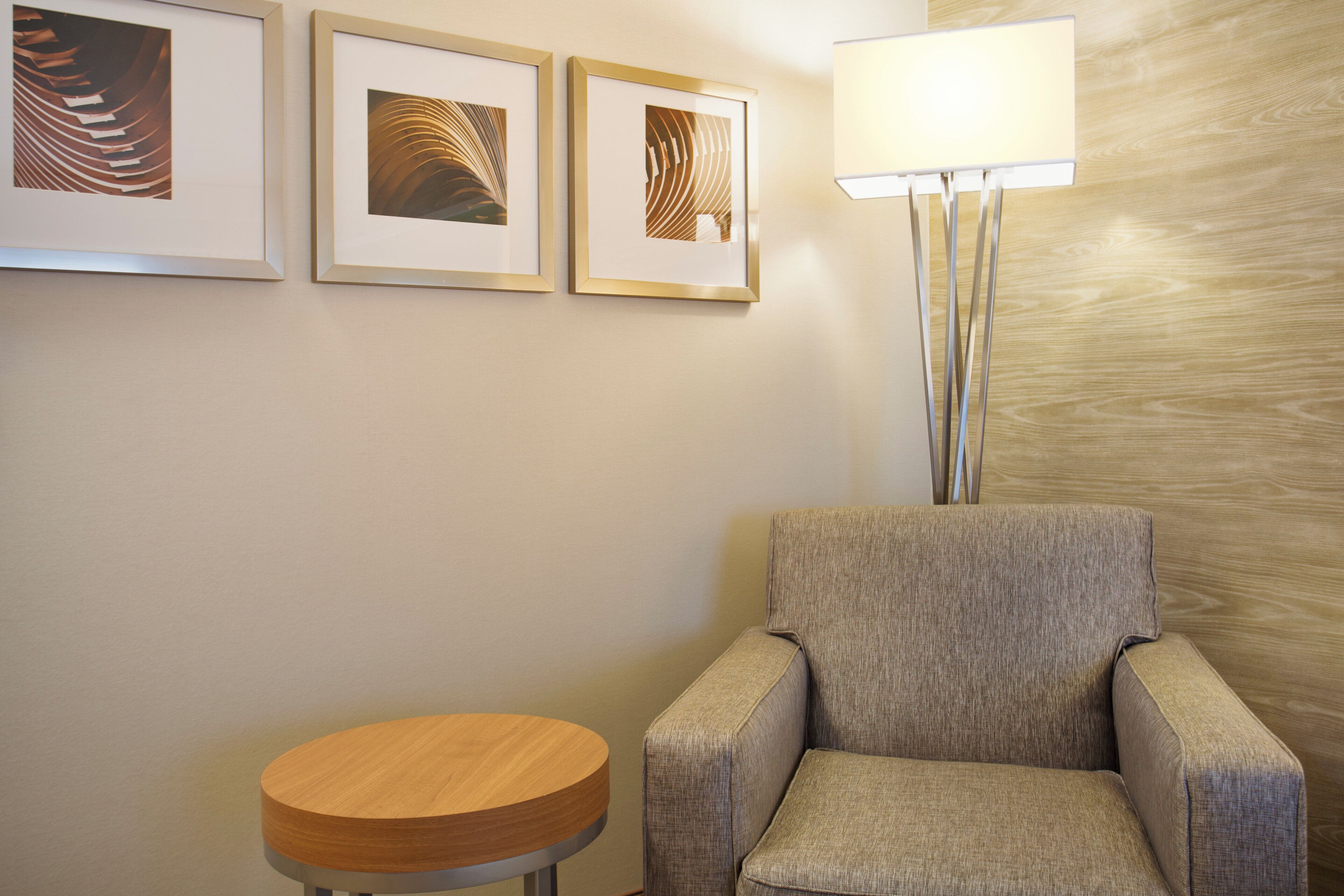 A hotel room with a grey armchair, a round wooden table, and three framed artworks on the wall.