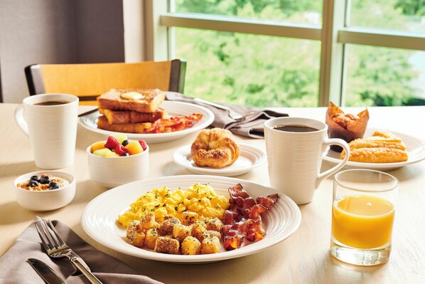 Una mesa de desayuno con platos de huevos revueltos, tocino, tostadas y pasteles, junto con tazas de café y jugo.