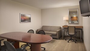 Ein Hotelzimmer mit Konferenztisch, zwei Stühlen, einer Couch, einem Schreibtisch mit Computer, einem Fernseher und einem gerahmten Bild an der Wand.