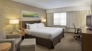 Ein Hotelzimmer mit einem großen Bett, einem Schreibtisch mit Fernseher, einem Sessel, einer Lampe und einem Gemälde an der Wand.