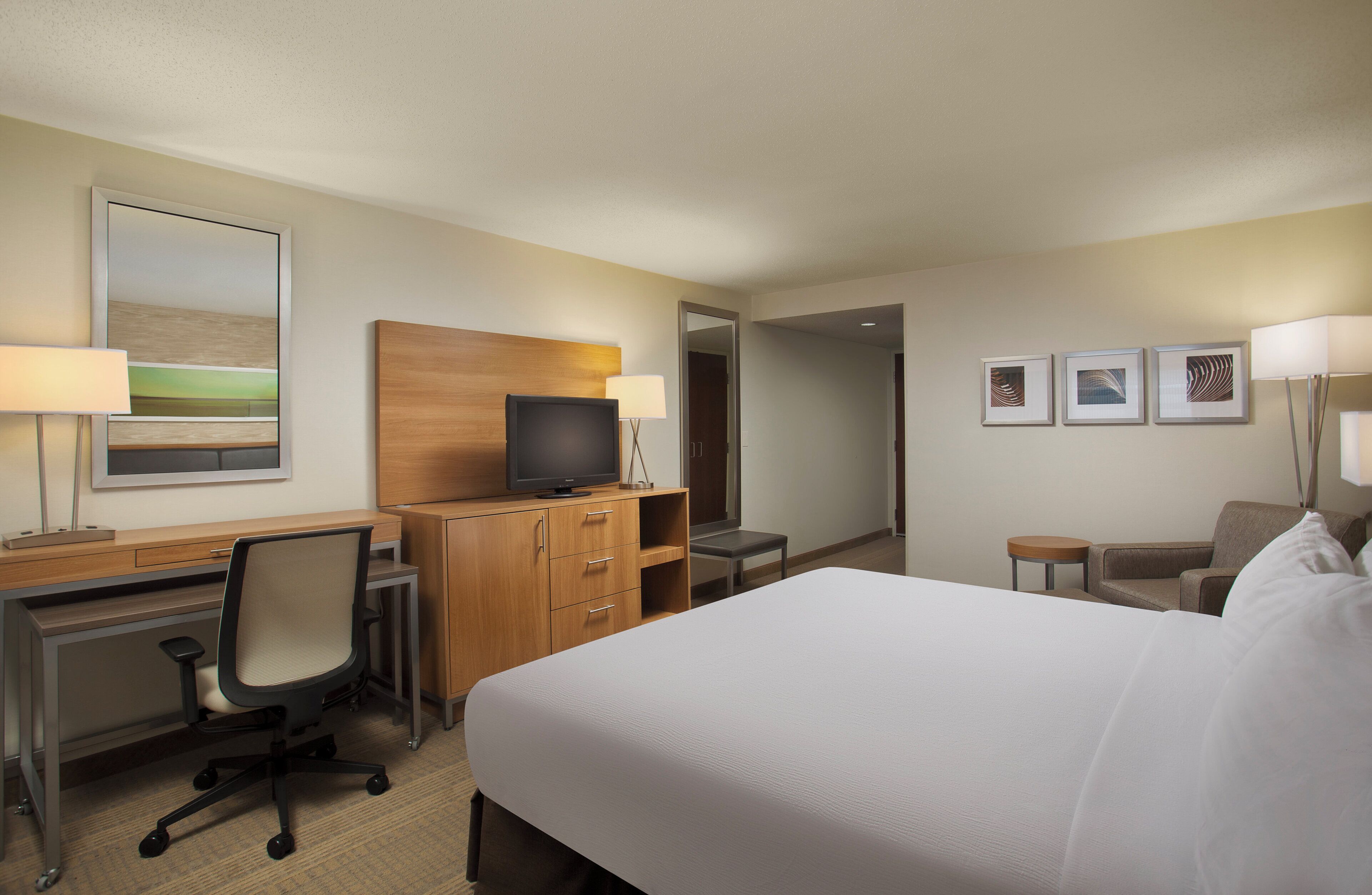 A hotel room with a bed, desk, chair, television, and artwork on the walls.