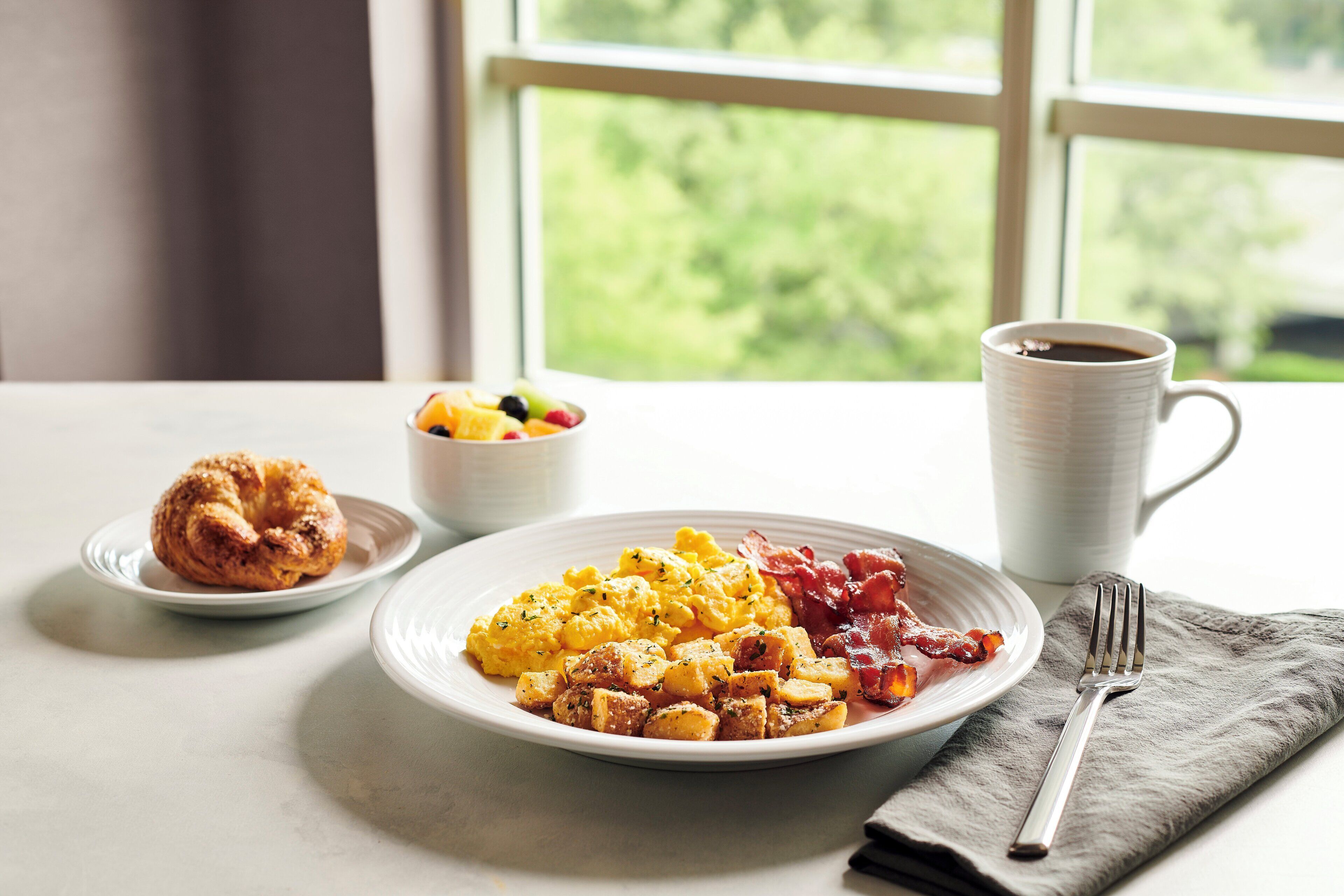 Un desayuno con huevos revueltos, tocino, un croissant y un bol de fruta sobre una mesa junto a una ventana.