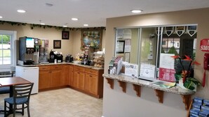 A kitchen with wooden cabinets, a coffee machine, and a refrigerator. There is a window with a view of a parking lot. - Scottish Inn Sturbridge (Sturbridge)