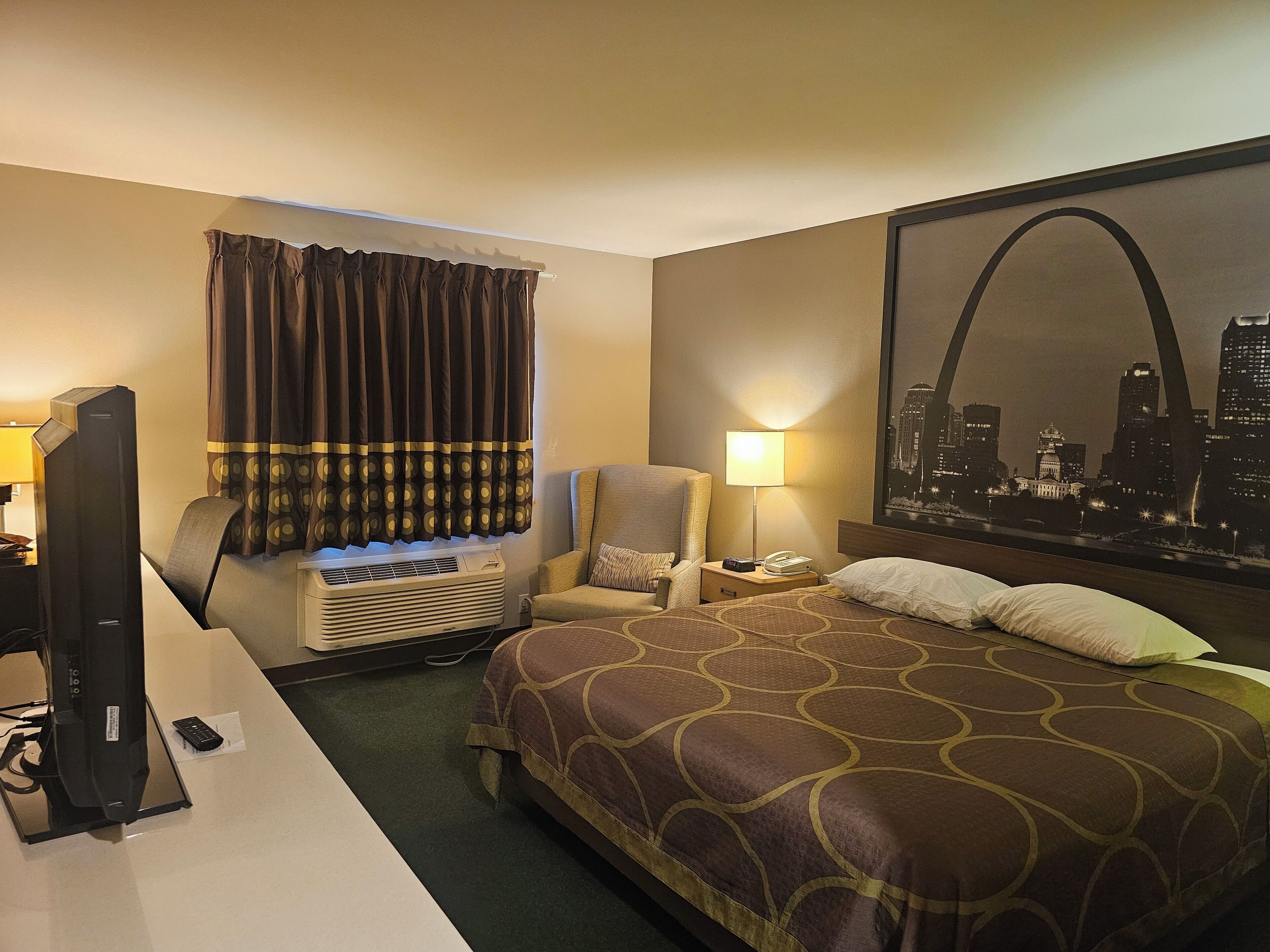 A hotel room with a bed, a chair, a desk with a TV, a lamp, and a large cityscape mural on the wall.