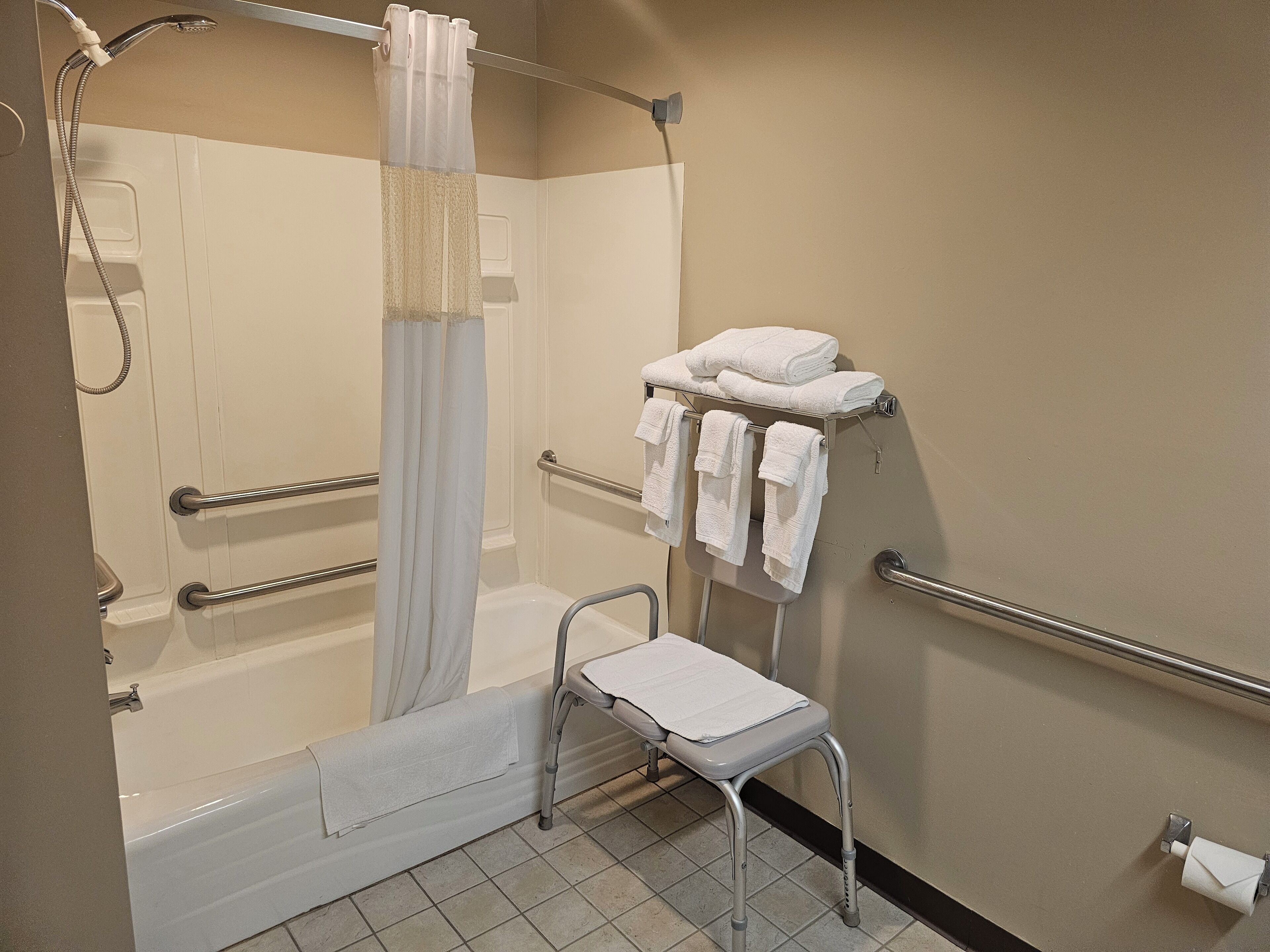 Un baño con ducha, barras de apoyo, un asiento accesible para silla de ruedas y un perchero para toallas.