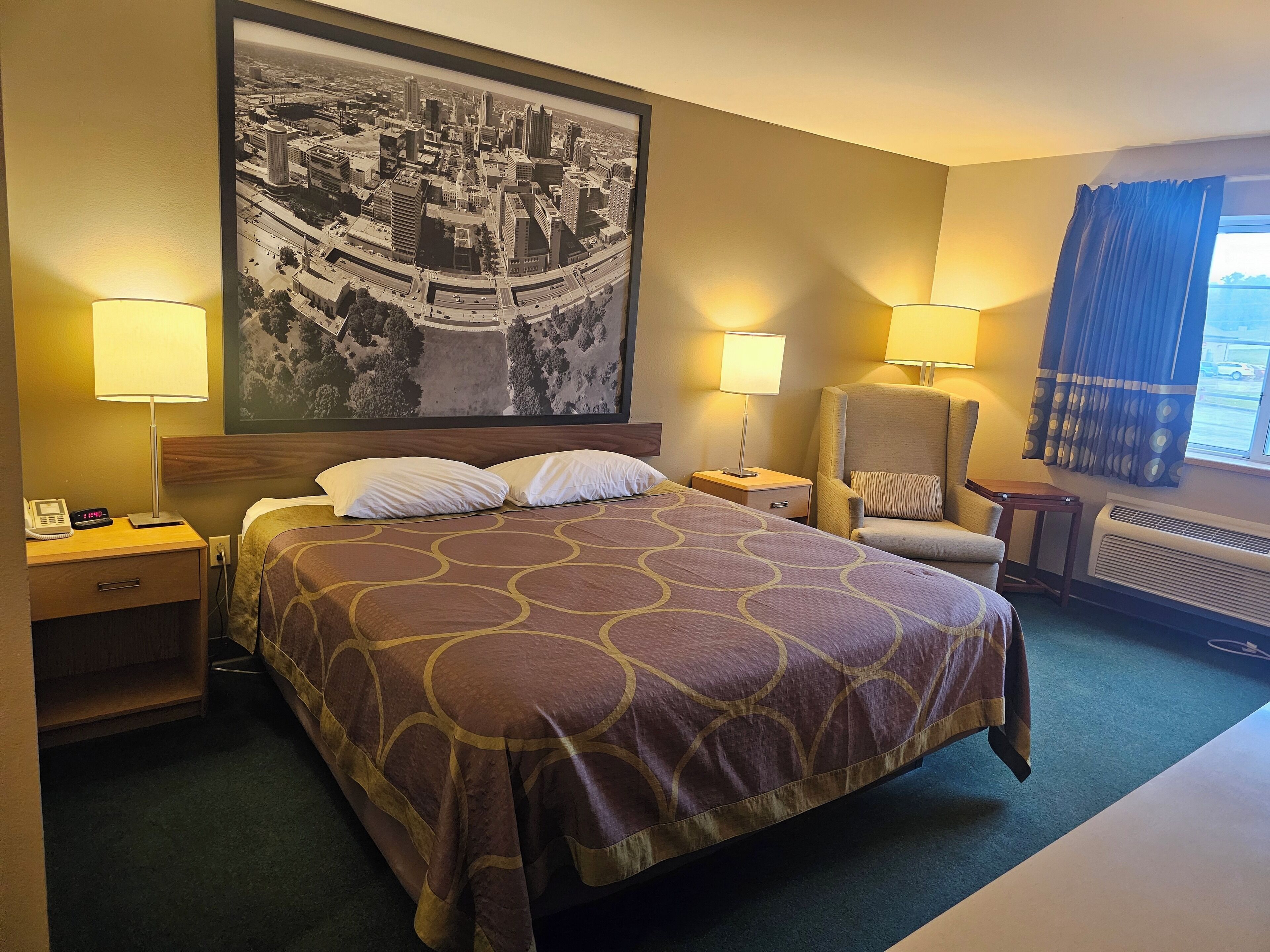Hotelkamer met een groot bed, twee nachtkastjes, een stoel, een groot raam met gordijnen en een stadsgezichtschilderij aan de muur.