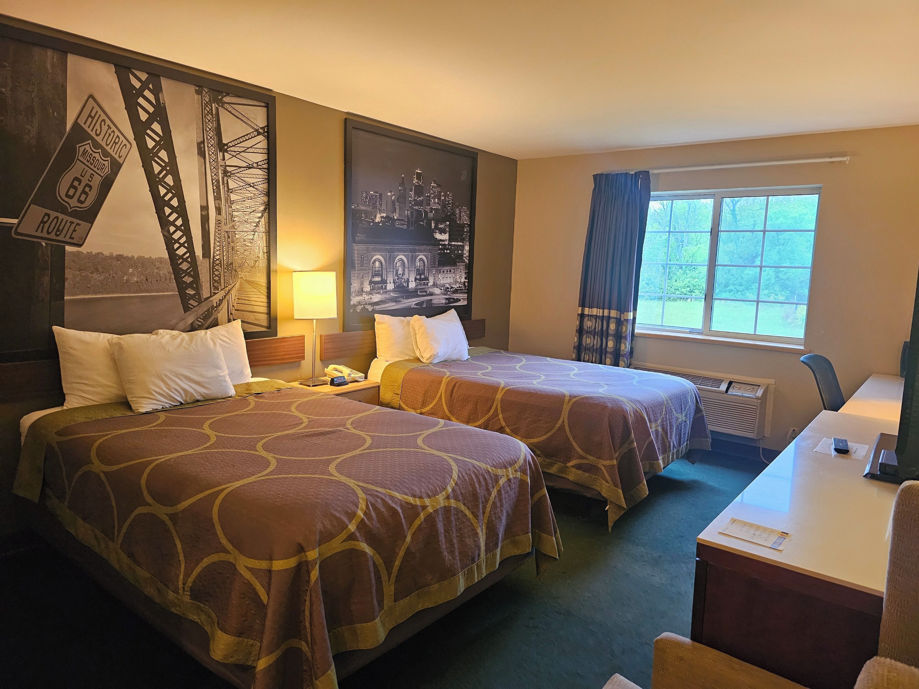 A hotel room with two beds, a desk, a window with curtains, and wall art including a cityscape and a historic route sign.