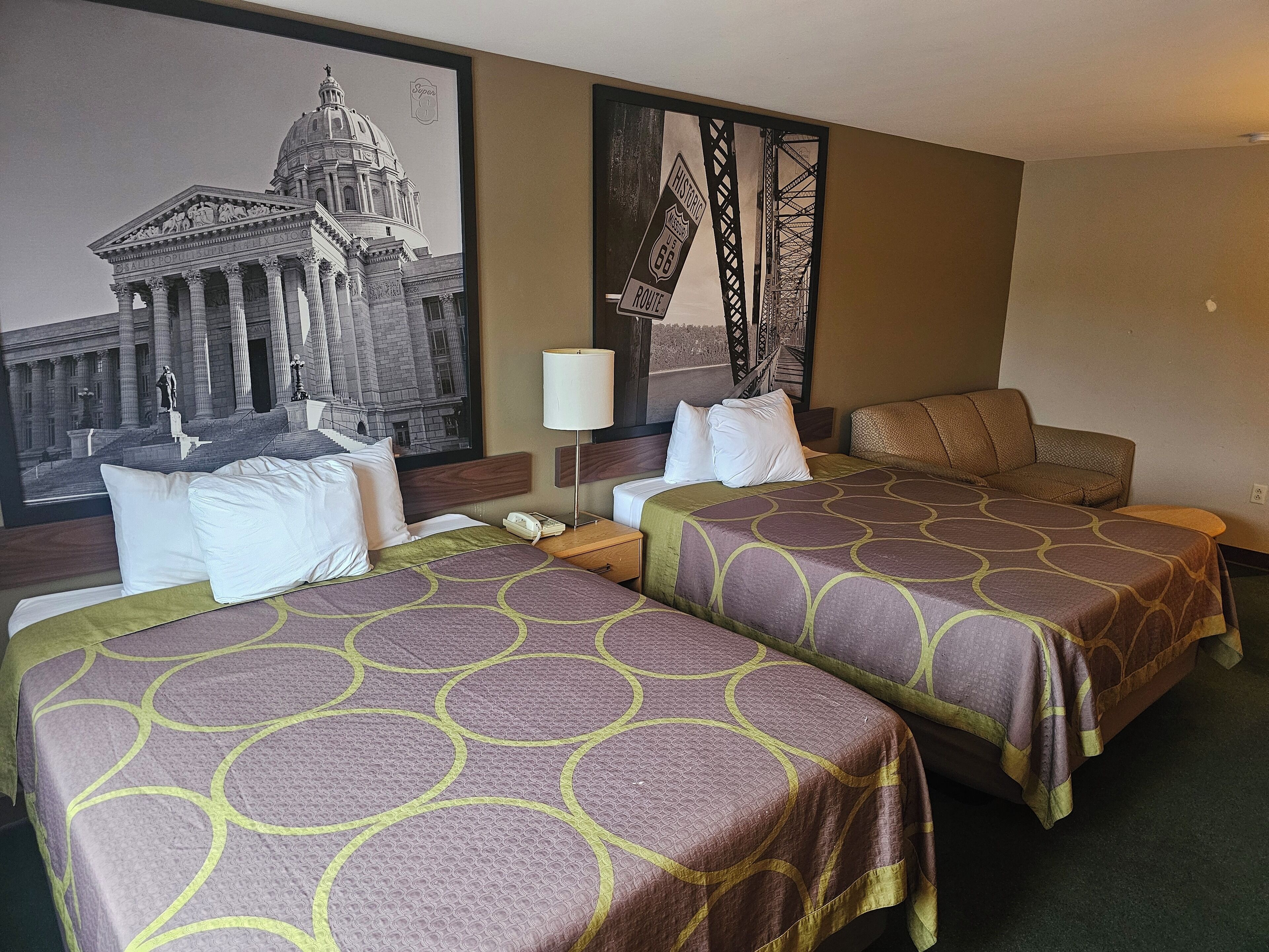 Ein Hotelzimmer mit zwei Betten, einem Nachttisch, einer Lampe und einem großen gerahmten Bild eines historischen Gebäudes.