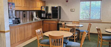A restaurant with wooden cabinets, a coffee machine, and a refrigerator. There are several wooden tables and chairs.