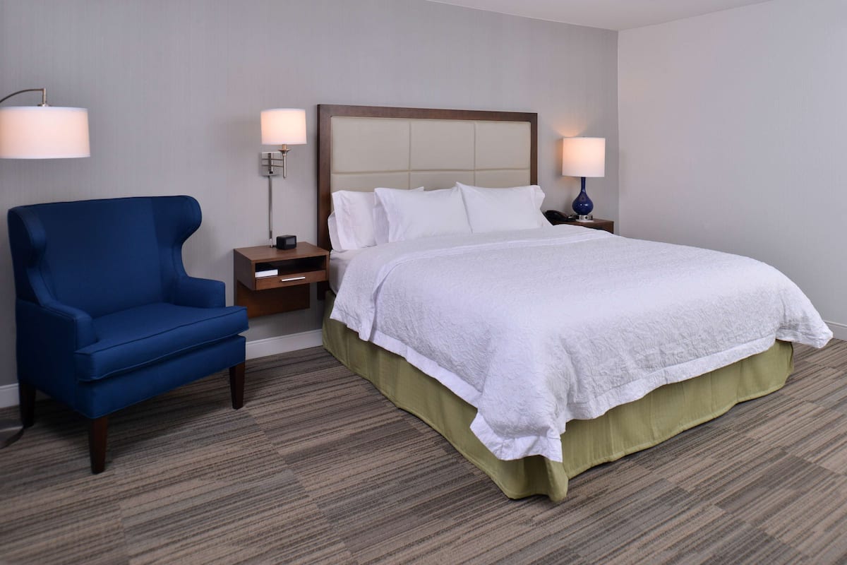 A hotel room with a bed, a blue armchair, two bedside tables with lamps, and a carpeted floor.