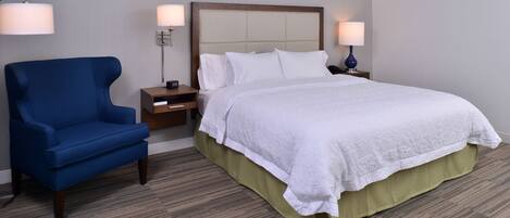 Una habitación de hotel con una cama, un sillón azul, dos mesitas de noche con lámparas y suelo alfombrado.