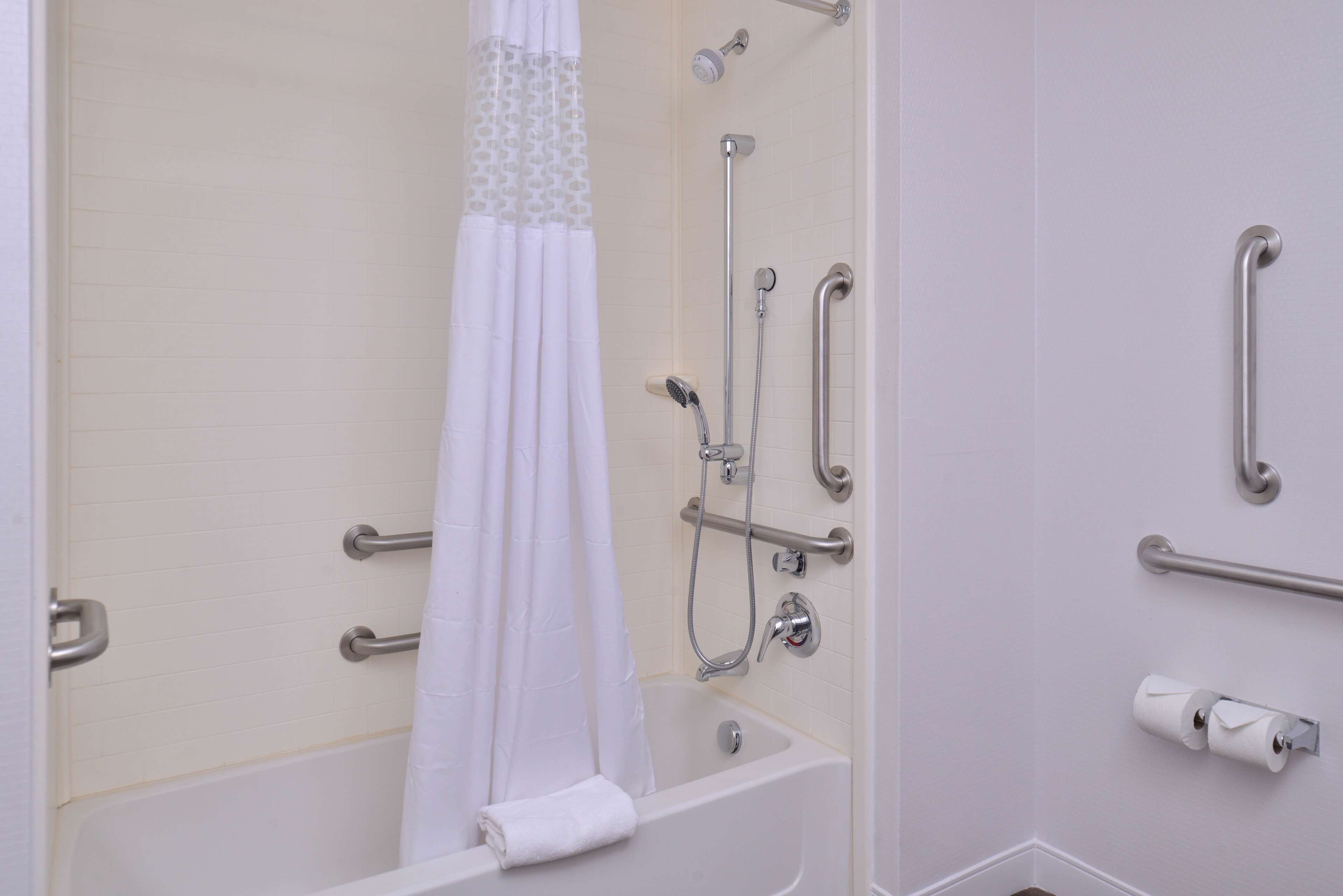 A bathroom with a bathtub, shower, grab bars, and a toilet paper holder.