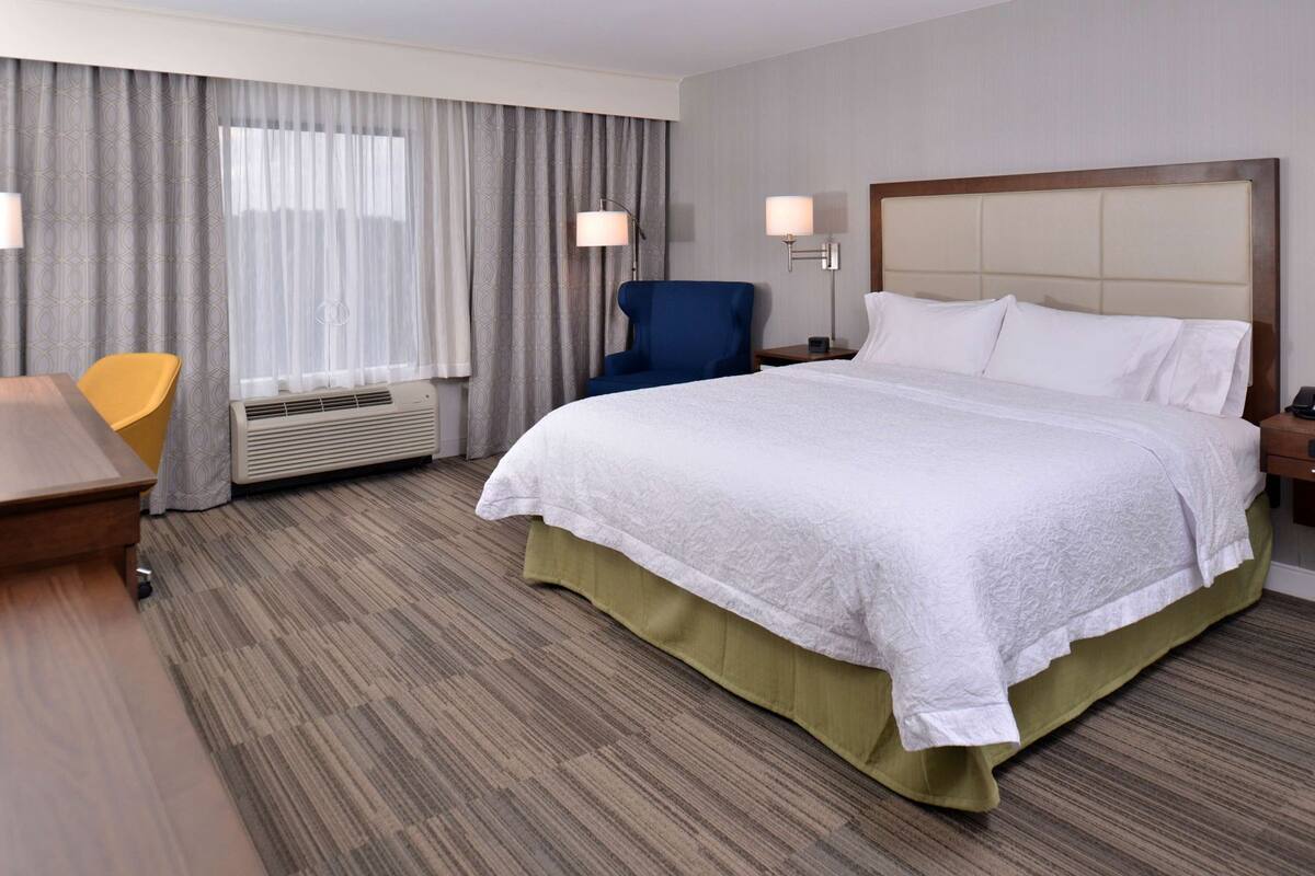 Quarto de hotel com cama, escrivaninha, cadeira, abajur e janela com cortinas.