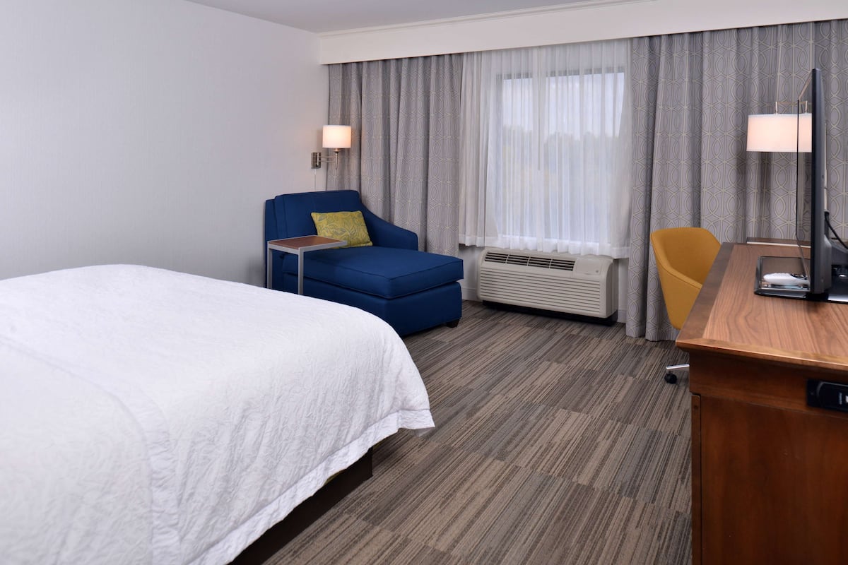 A hotel room with a bed, a blue armchair, a wooden desk, and a television.