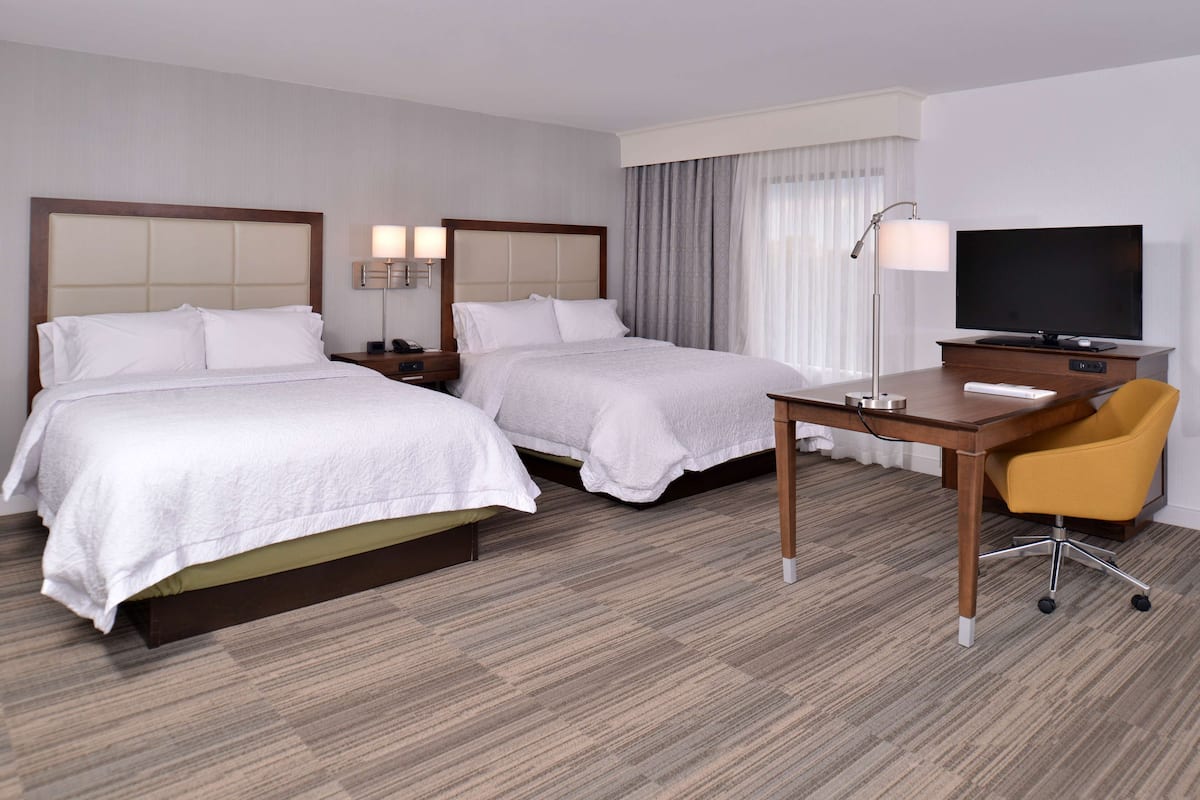 A hotel room with two beds, a desk, a chair, a television, and a lamp.