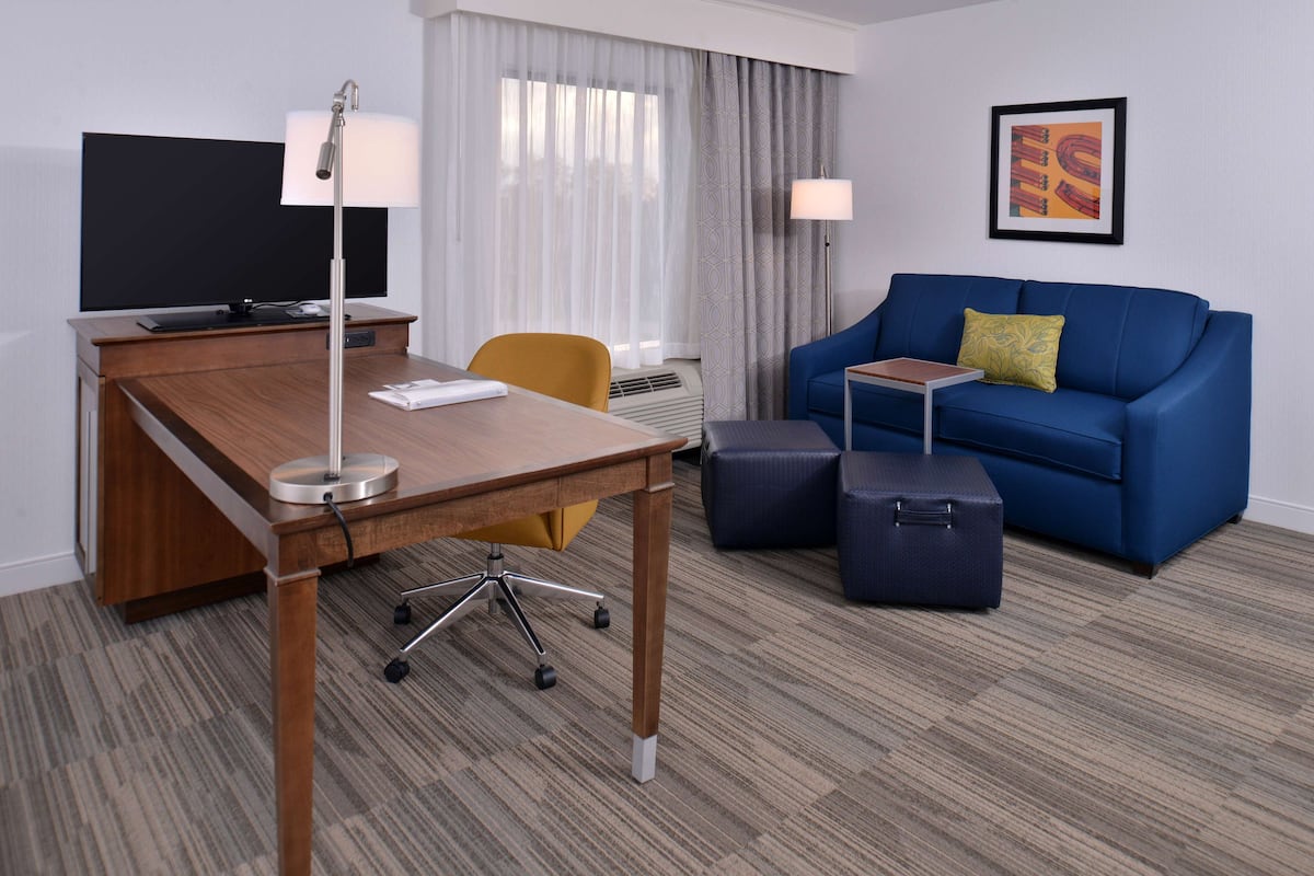 A hotel room with a wooden desk, a blue sofa, a yellow chair, a black TV, a lamp, a suitcase, and a framed picture on the wall.