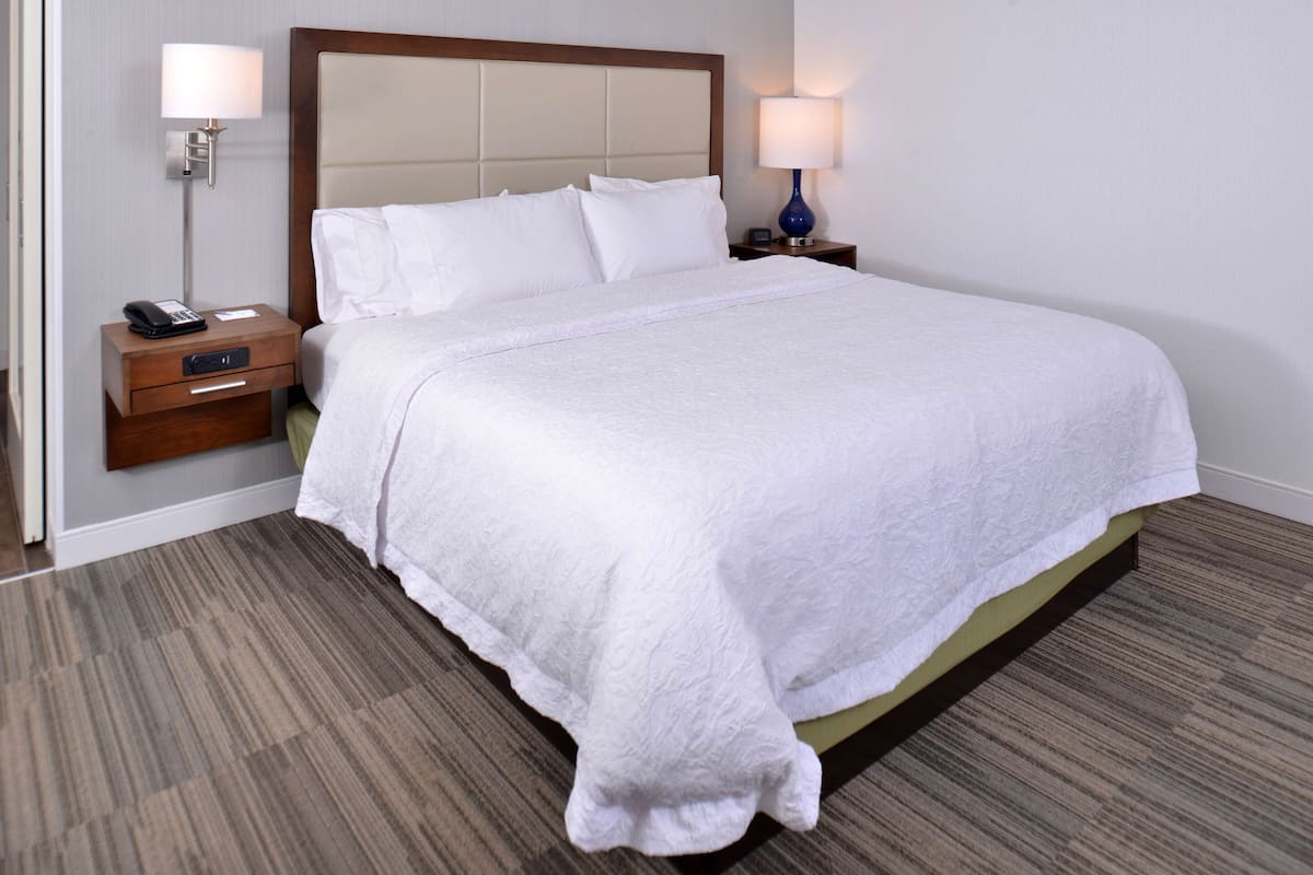 A neatly made bed with a white duvet, a wooden headboard, and a nightstand with a lamp and a phone.
