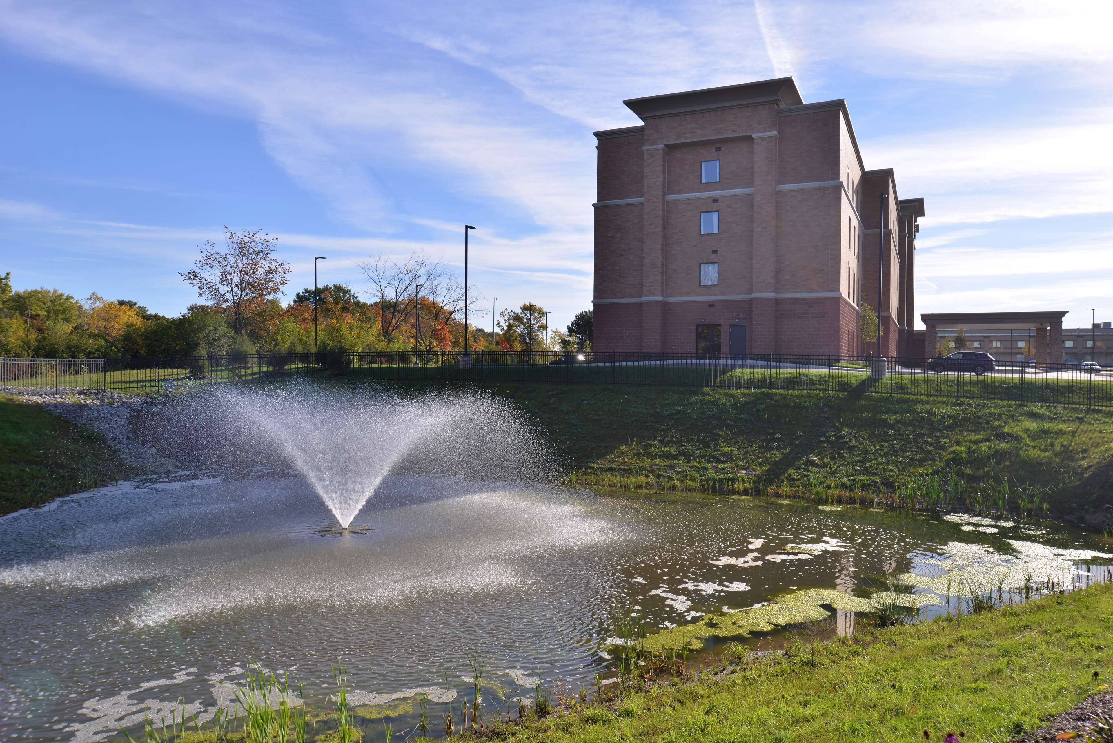 A building with a brick facade and multiple windows, a fountain in a pond with lily pads, and a clear sky.