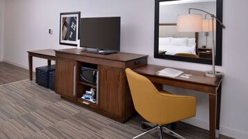 A hotel room with a wooden desk, a yellow chair, a flat-screen TV, a mirror, and a lamp.