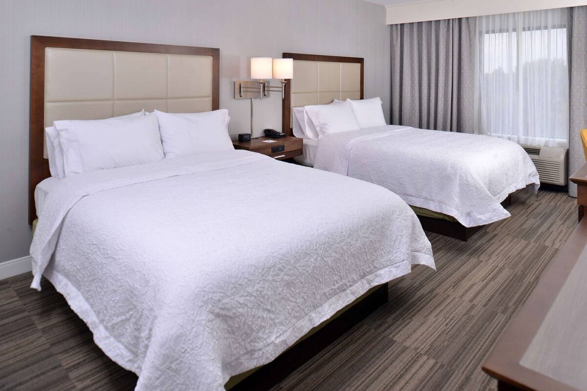 Quarto de hotel com duas camas, uma mesinha de cabeceira com abajur e uma janela com cortinas.