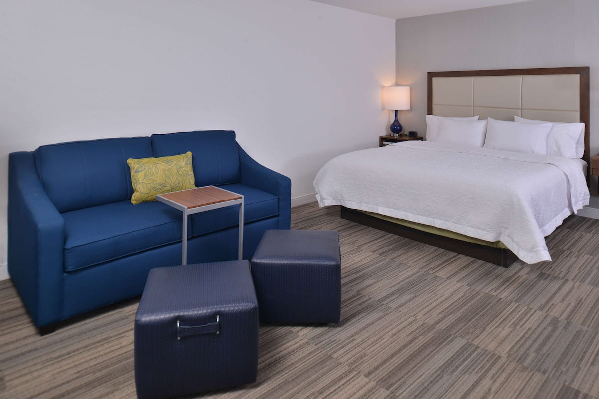 A hotel room with a blue sofa, a small wooden table, two blue ottomans, a large bed with white bedding, and a nightstand with a lamp.