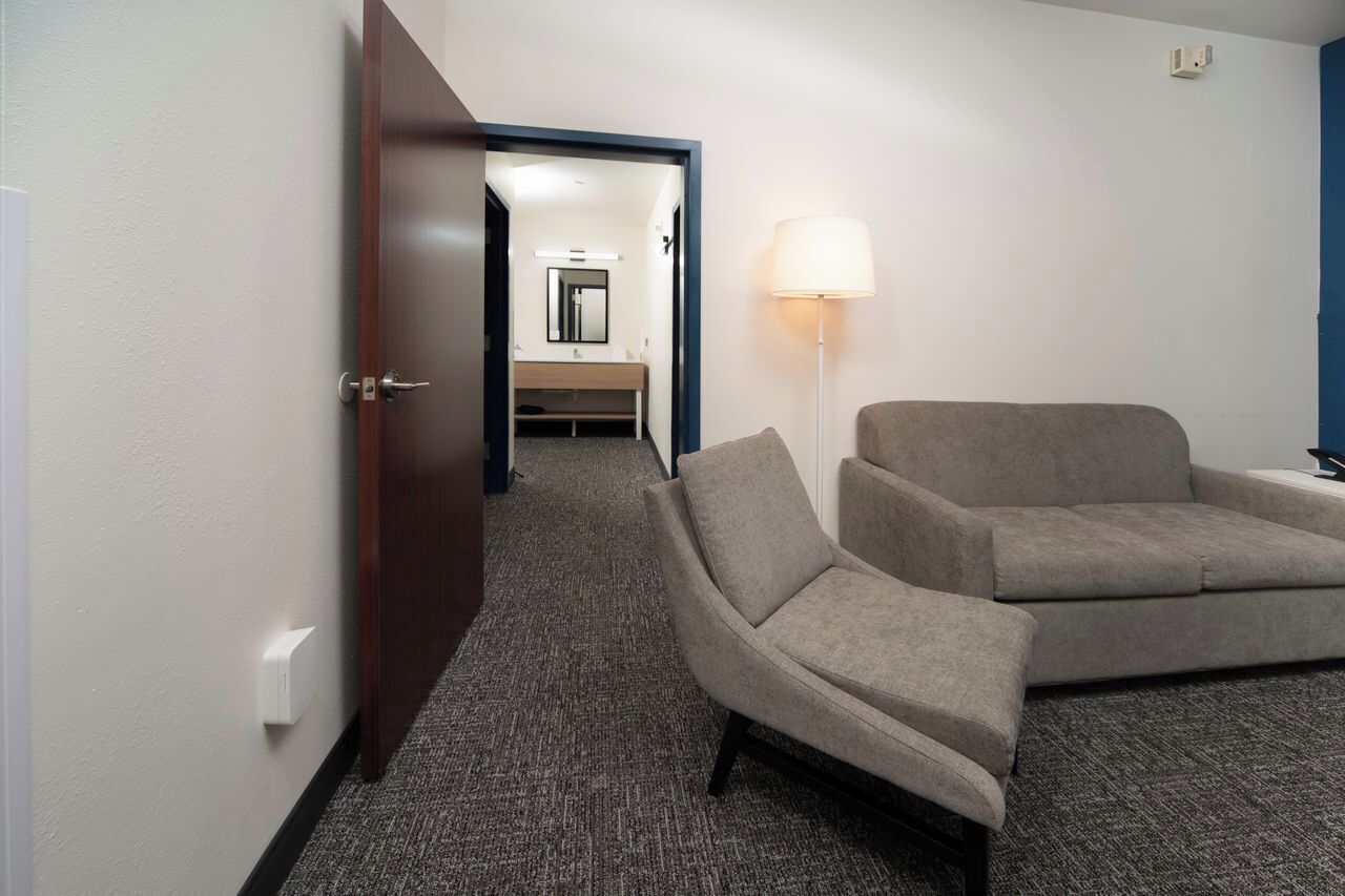 A hotel room with a dark wooden door, a grey carpet, a grey sofa, a floor lamp, and a bathroom visible through an open door.