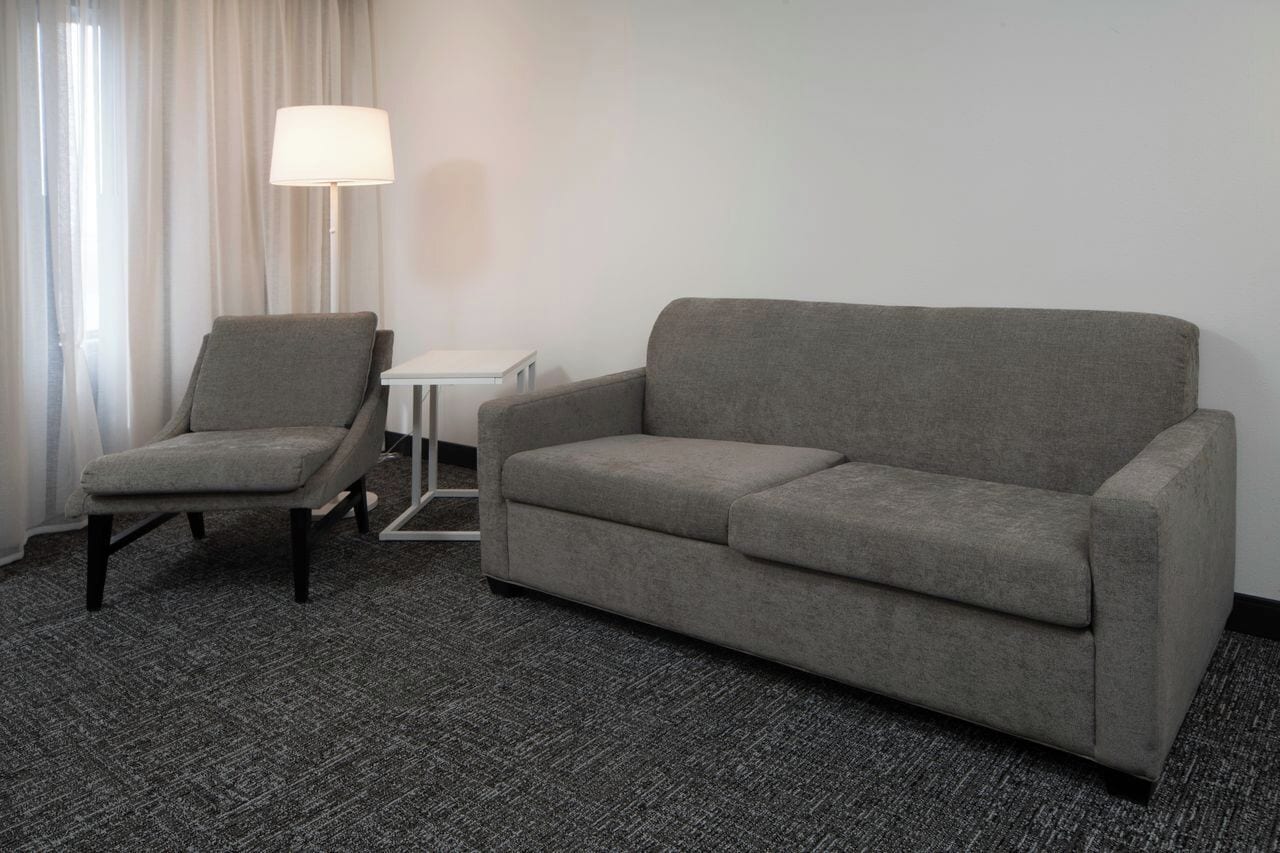 Habitación de hotel con un sillón gris, un sofá gris, una mesita auxiliar blanca y una lámpara de pie.