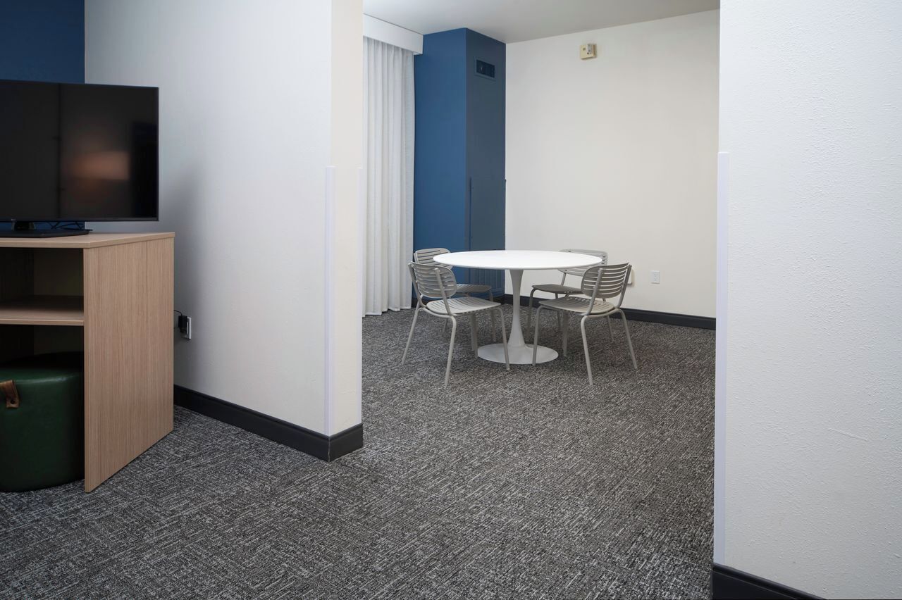 A hotel room with a flat-screen TV, a small wooden cabinet, a green ottoman, a round table with four chairs, and a blue accent wall.