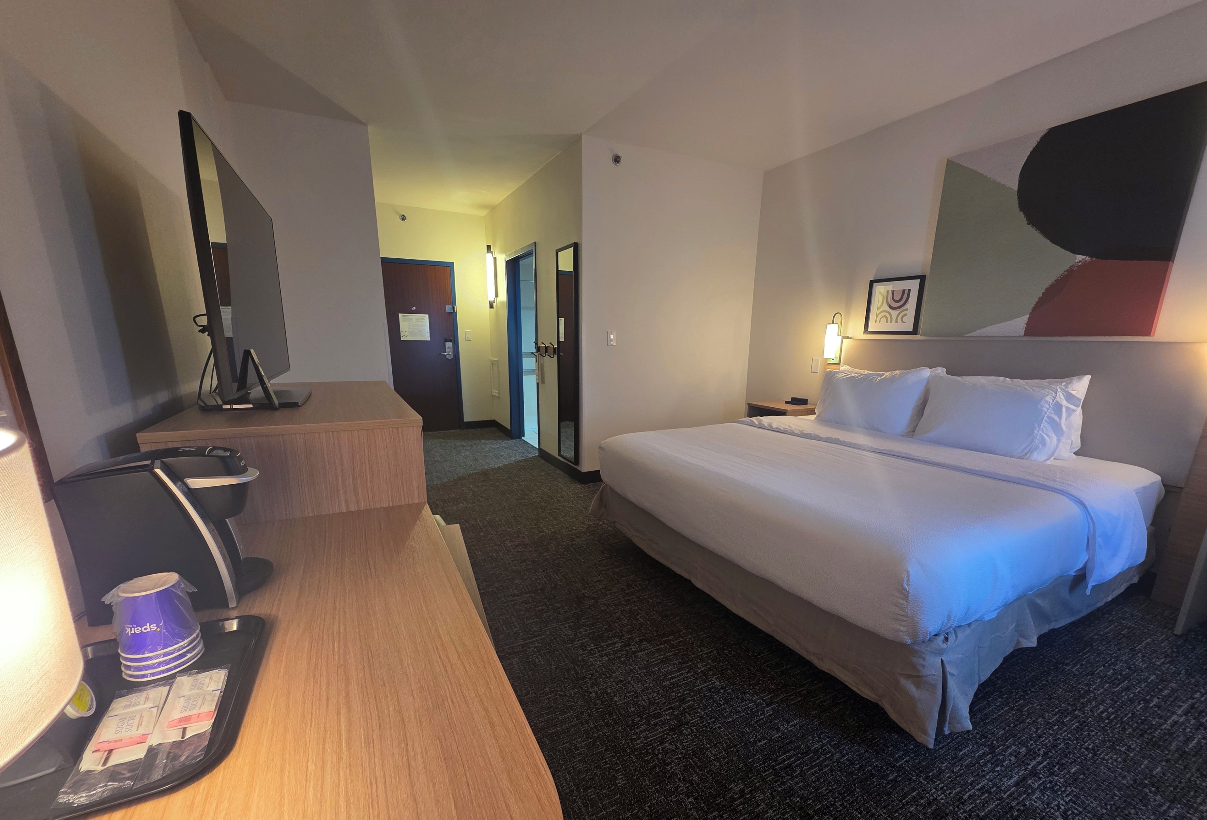 Habitación de hotel con una cama, un escritorio con una computadora, una silla y una pequeña mesa con una bolsa y una botella.