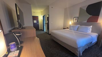 Habitación de hotel con una cama, un escritorio con una computadora, una silla y una pequeña mesa con una bolsa y una botella.