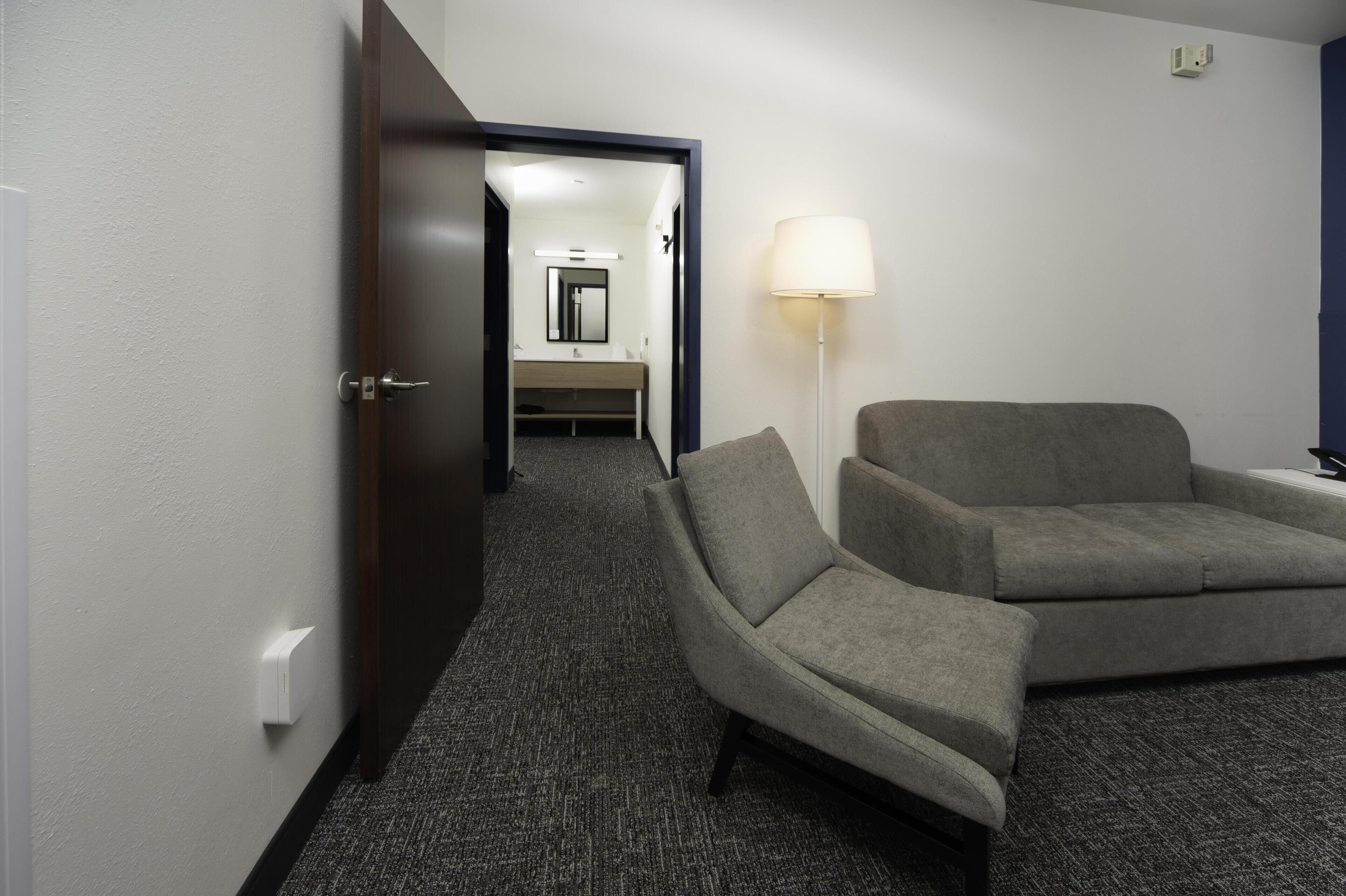 Habitación de hotel con un sofá gris, una lámpara de pie y vistas a un baño con espejo y lavabo.