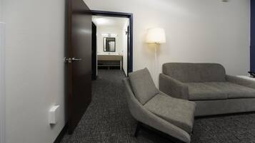 Habitación de hotel con un sofá gris, una lámpara de pie y vistas a un baño con espejo y lavabo.
