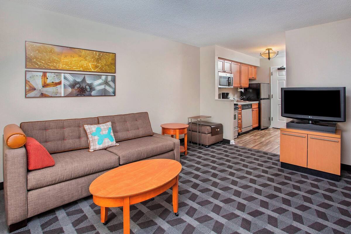 A living room with a sofa, coffee table, and artwork on the wall. A kitchen is visible in the background with cabinets and appliances.