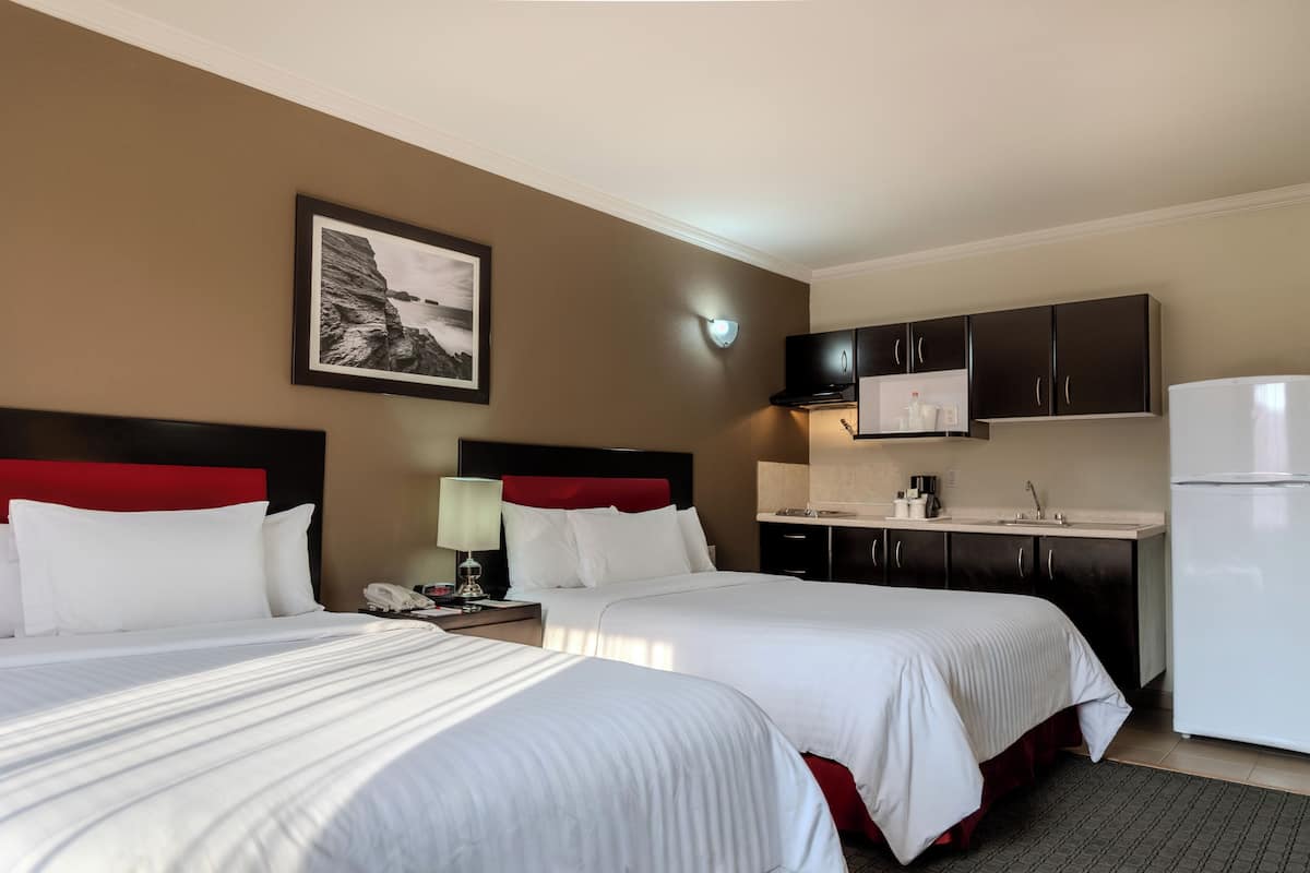 A hotel room with two beds, a kitchenette, a framed picture on the wall, and a nightstand with a lamp.