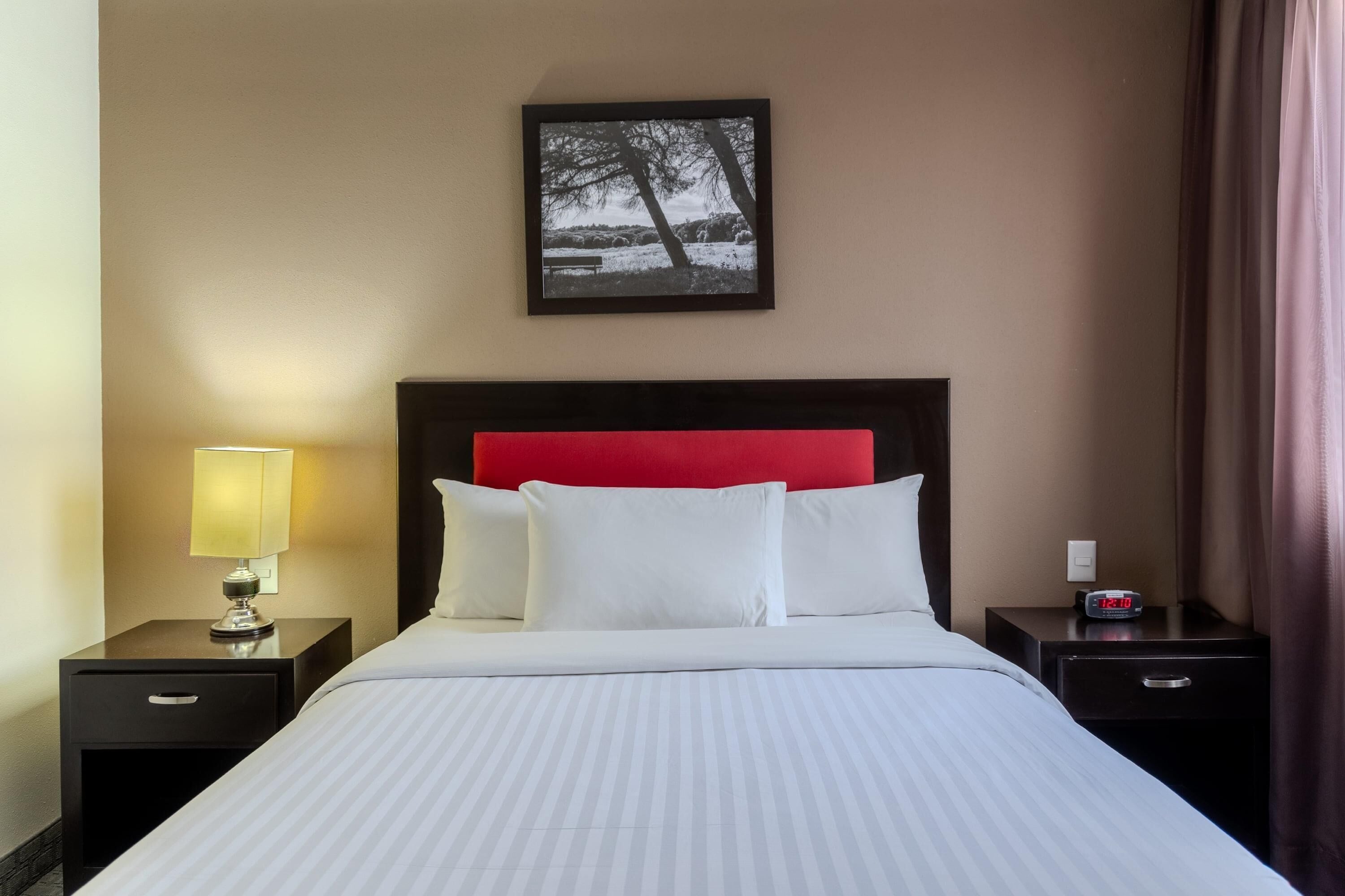 A hotel room with a bed, a nightstand with a lamp, a framed picture on the wall, and a clock.