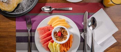 Un plato de desayuno con tortitas, fruta, granola, una taza de café y un vaso de zumo de naranja sobre una mesa.