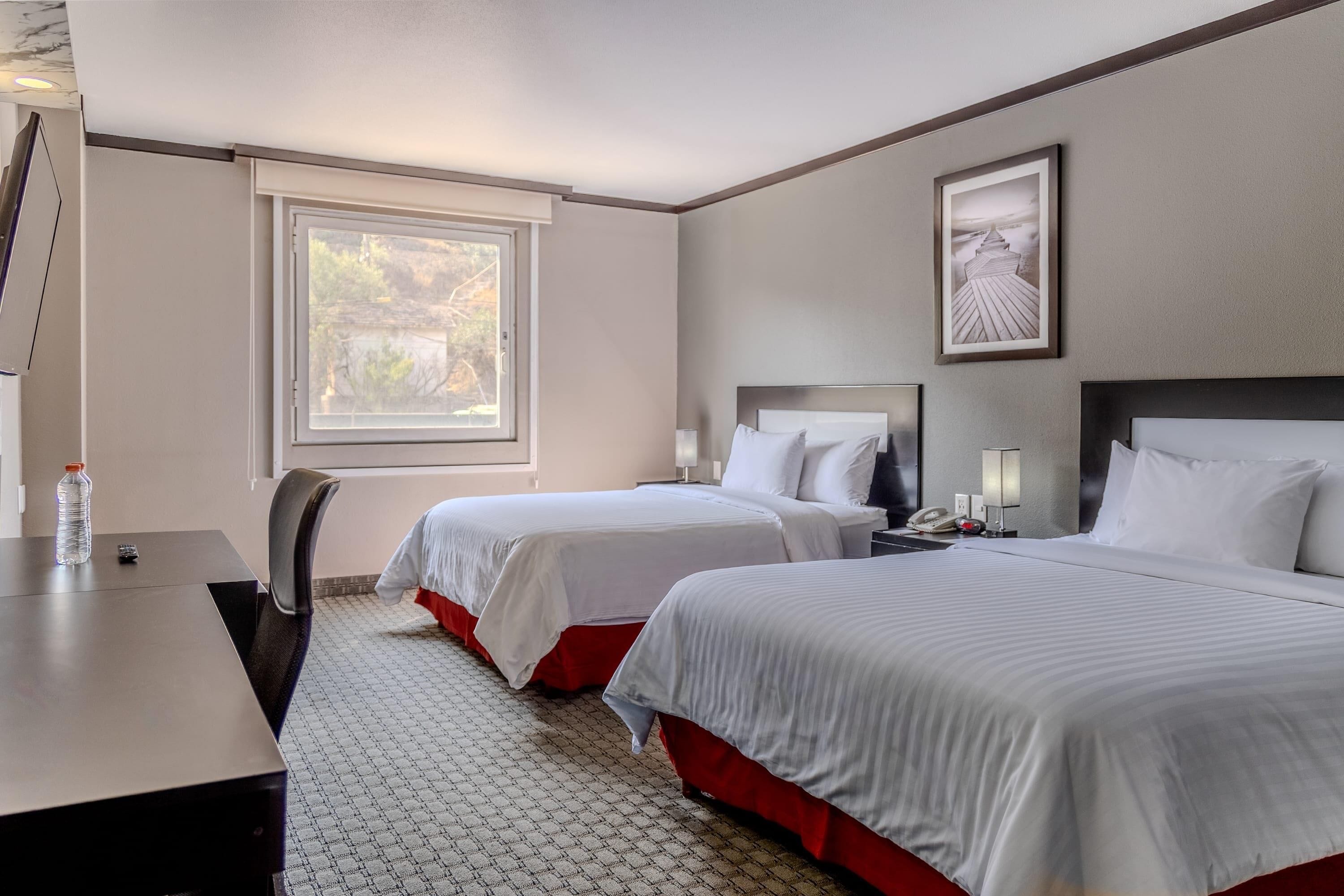 Une chambre d’hôtel avec deux lits, un bureau, une chaise, une fenêtre et un tableau encadré au mur.