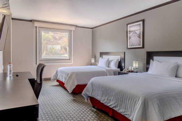 A hotel room with two beds, a desk, a chair, a window, and a framed picture on the wall.