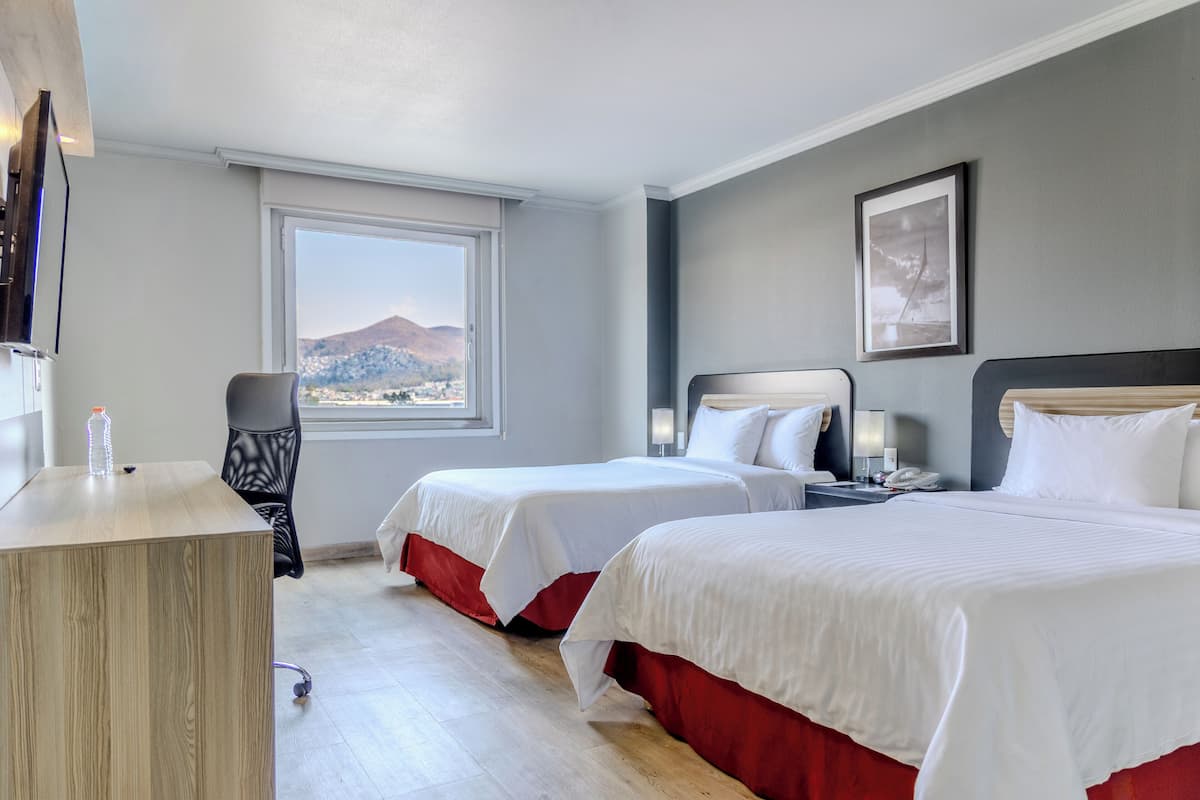 A hotel room with two beds, a desk, a chair, a window with a mountain view, and a framed picture on the wall.
