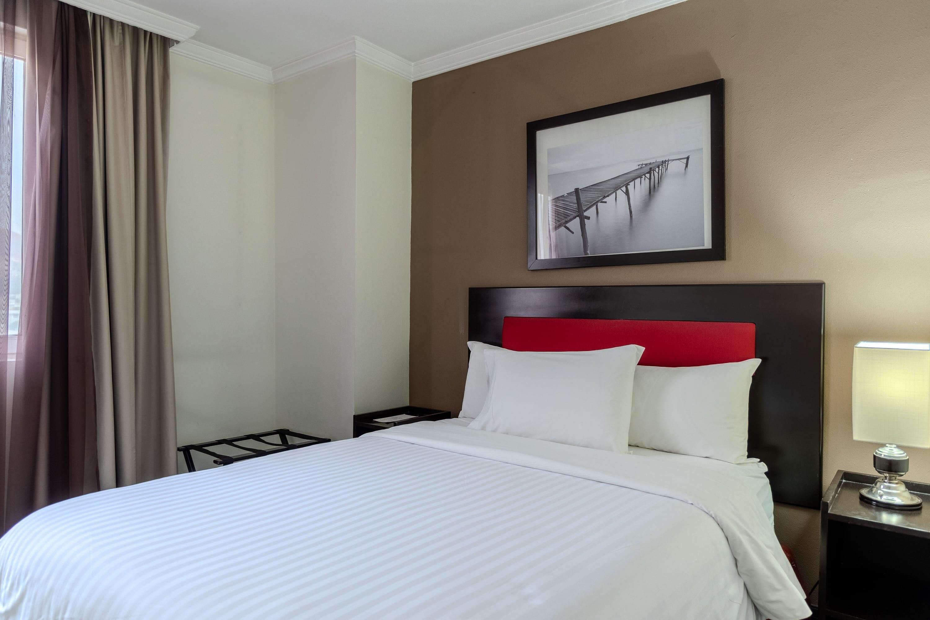 A hotel room with a bed, a nightstand, a lamp, a framed picture on the wall, and a window with curtains.
