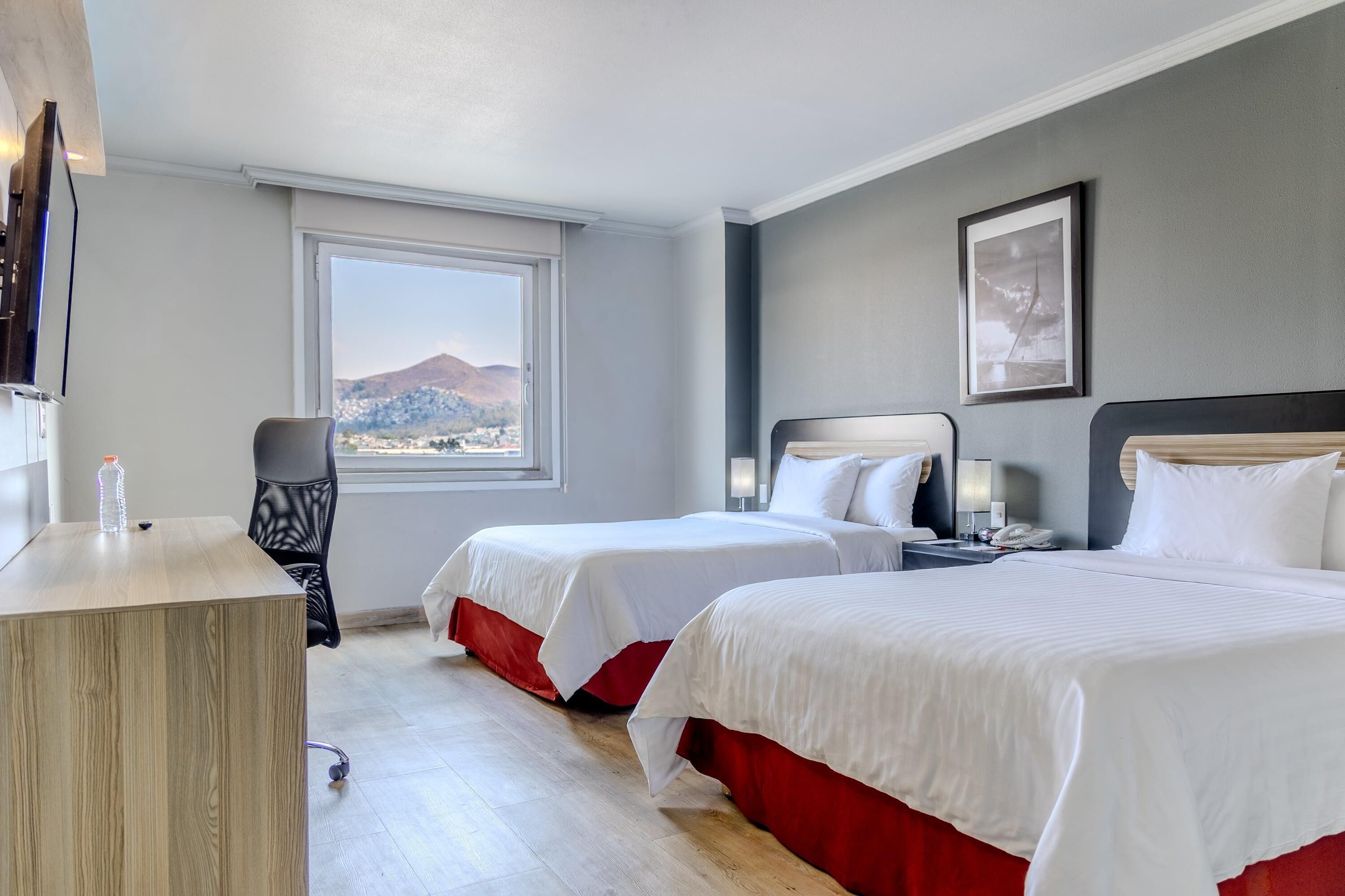 A hotel room with two beds, a desk, a chair, a window with a mountain view, and a framed picture on the wall.