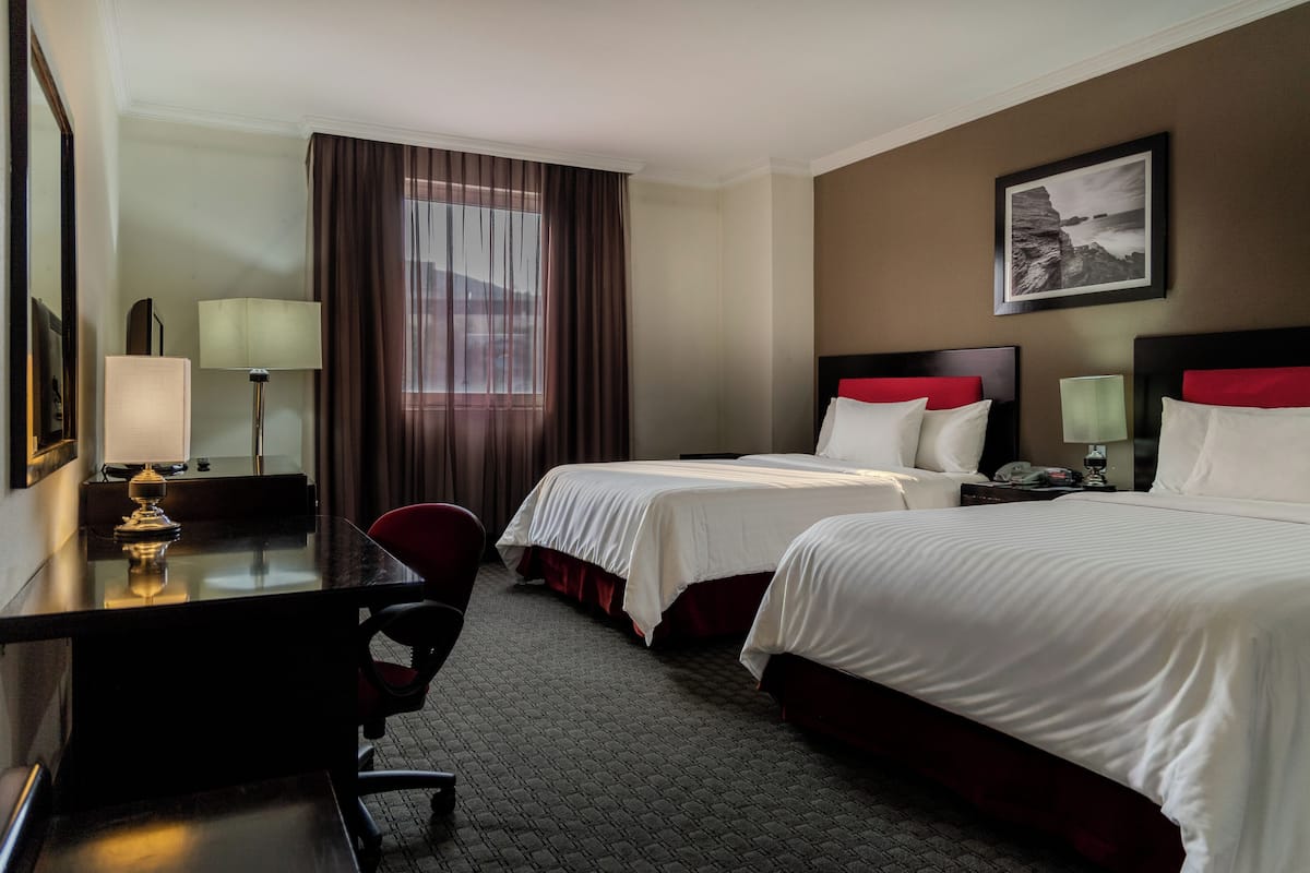 A hotel room with two beds, a desk, a chair, a lamp, and a framed picture on the wall.