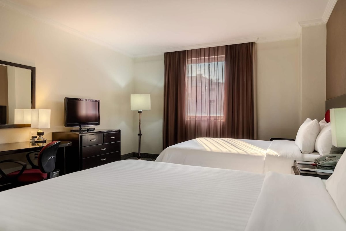 A hotel room with two beds, a desk with a television, a lamp, and a large window with curtains.