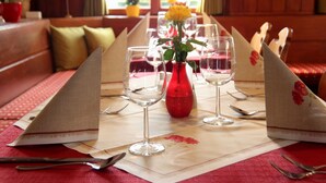 Una mesa de comedor preparada con copas de vino, un florero rojo con una flor amarilla y un mantel rojo.