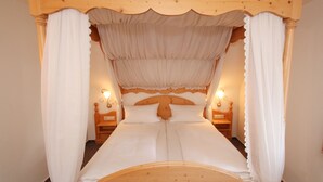Una cama con dosel y cuatro columnas, con ropa de cama blanca, cabecera de madera y dos mesitas de noche con lámparas.