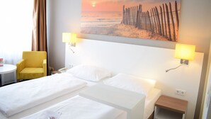 Una habitación de hotel con una cama, dos lámparas de noche, una mesita, una silla y un cuadro con una playa.