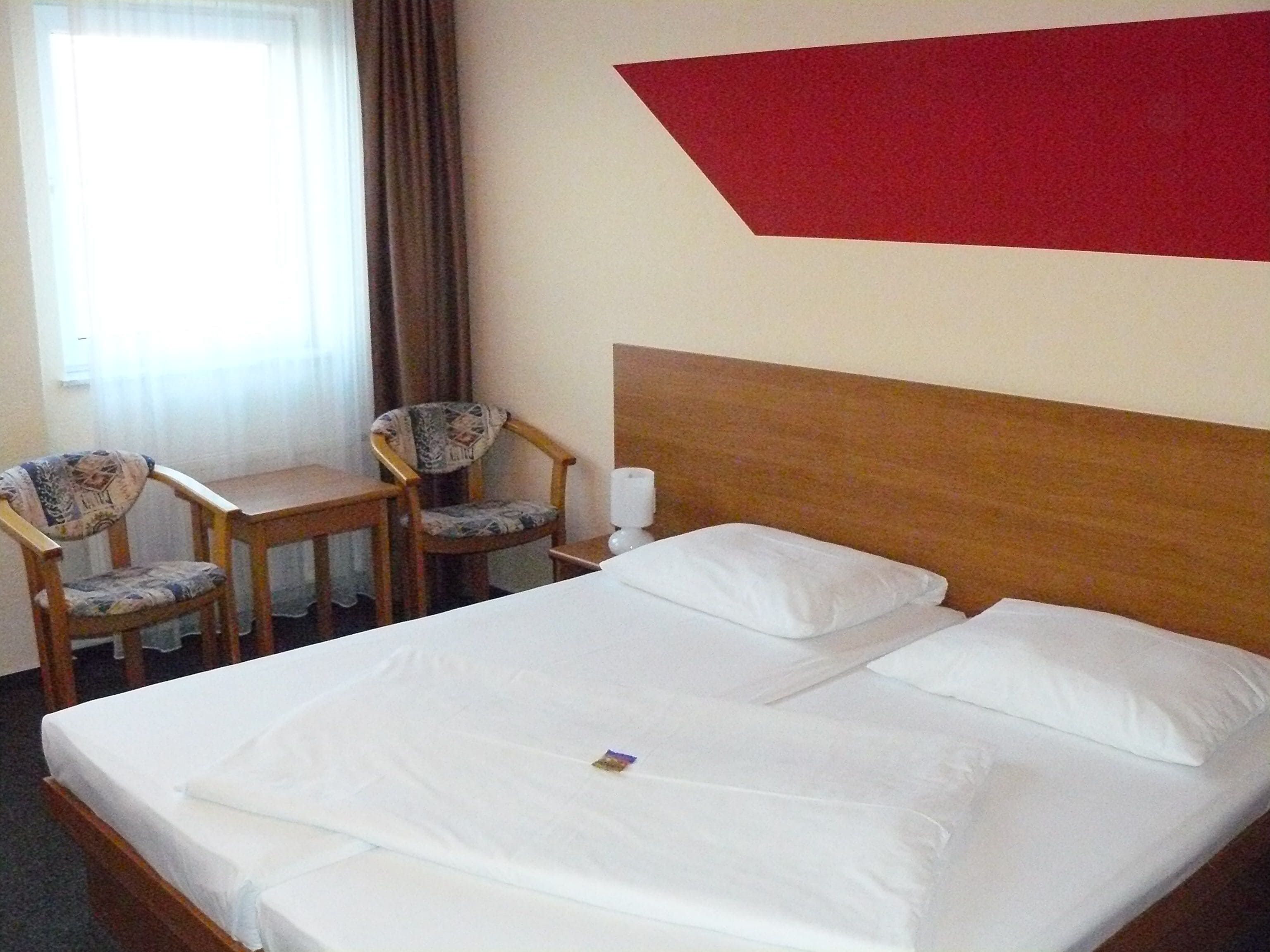 Habitación de hotel con cama, dos sillas, una mesita de noche y una pared roja decorativa.